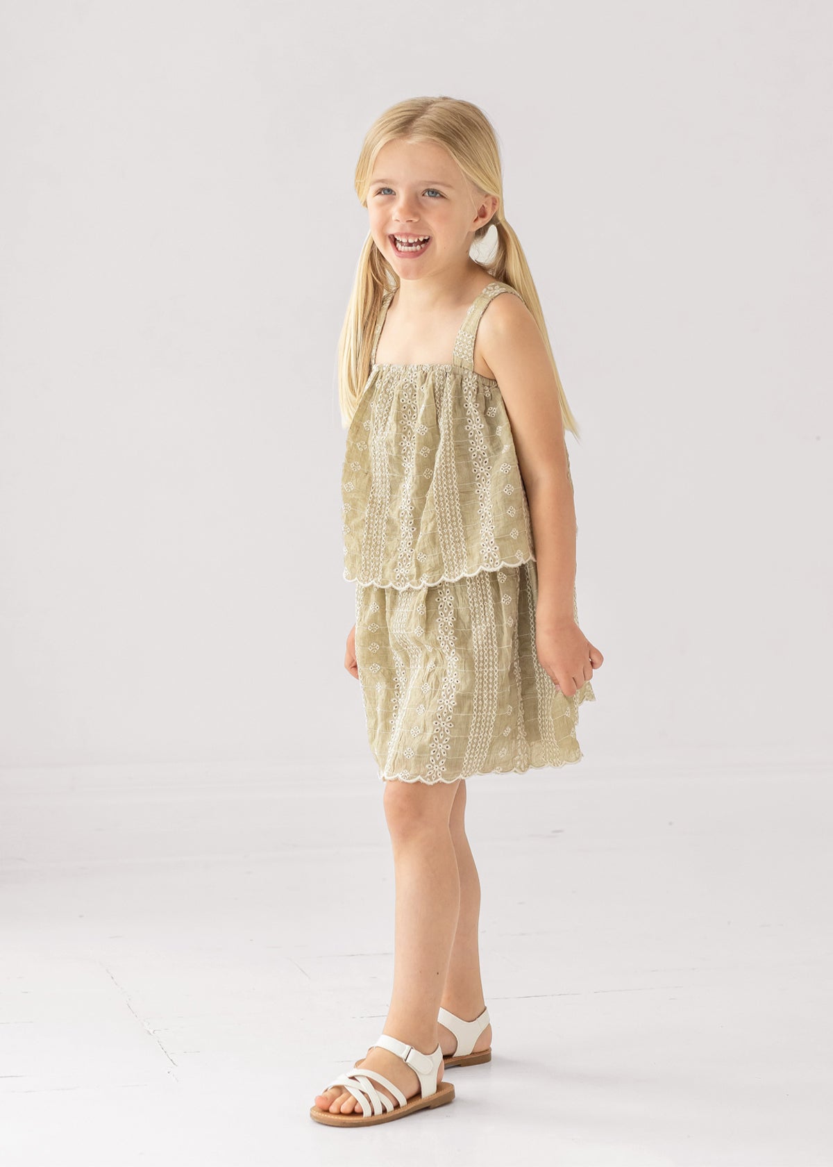 A young girl with blonde pigtails smiles in a Mabel and Honey Espresso Sleeveless Dress with embroidery and a scalloped hem, paired with white sandals. The background is plain and light-colored.