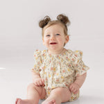 A smiling baby with light brown hair in two buns sits on the floor wearing the Mabel and Honey Audrey Flutter Sleeve Romper, featuring puffed sleeves and multicolored patterns, against a white background.