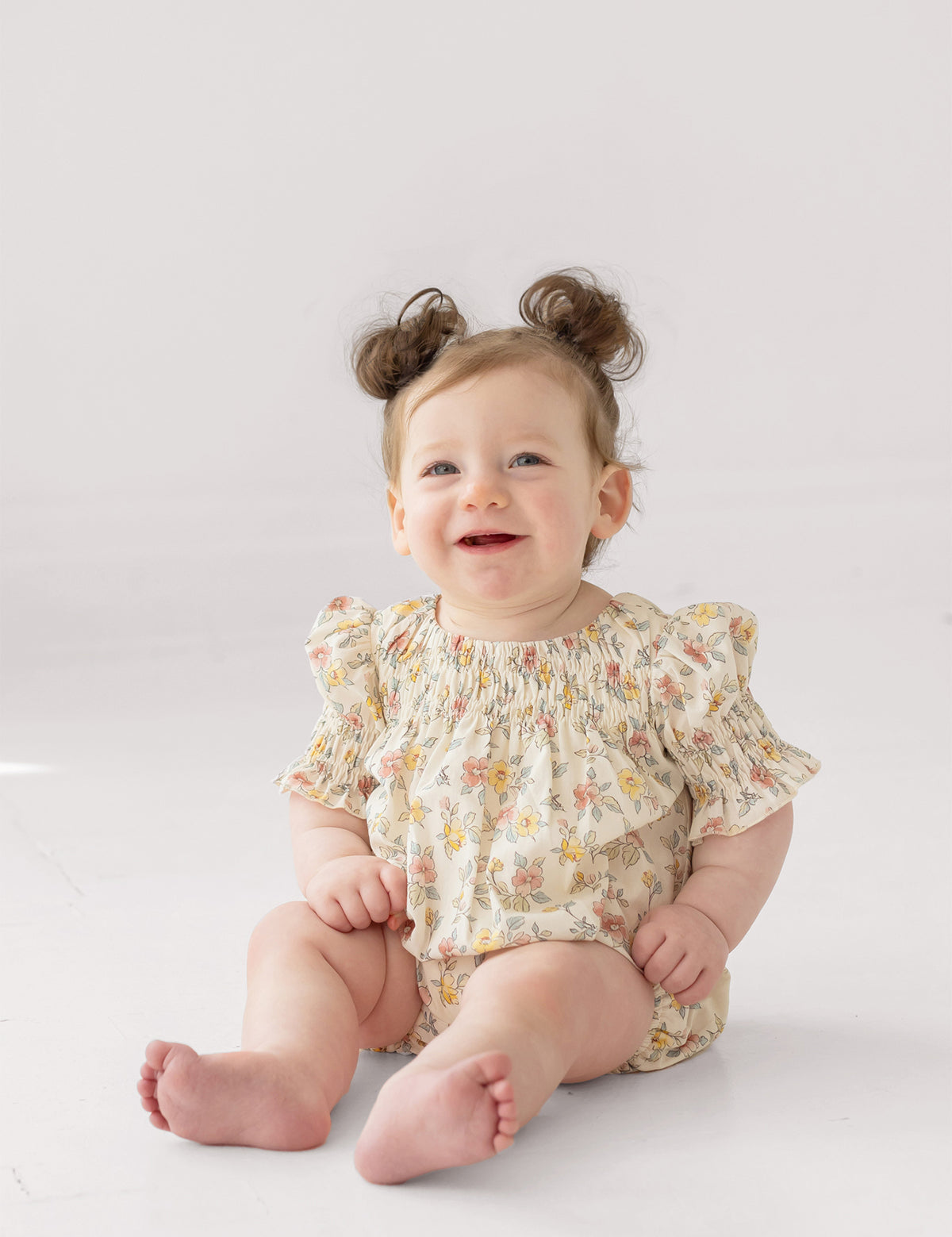 A smiling baby with light brown hair in two buns sits on the floor wearing the Mabel and Honey Audrey Flutter Sleeve Romper, featuring puffed sleeves and multicolored patterns, against a white background.