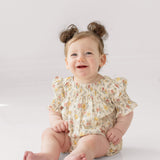 A smiling baby with light brown hair in two buns sits on the floor wearing the Mabel and Honey Audrey Flutter Sleeve Romper, featuring puffed sleeves and multicolored patterns, against a white background.