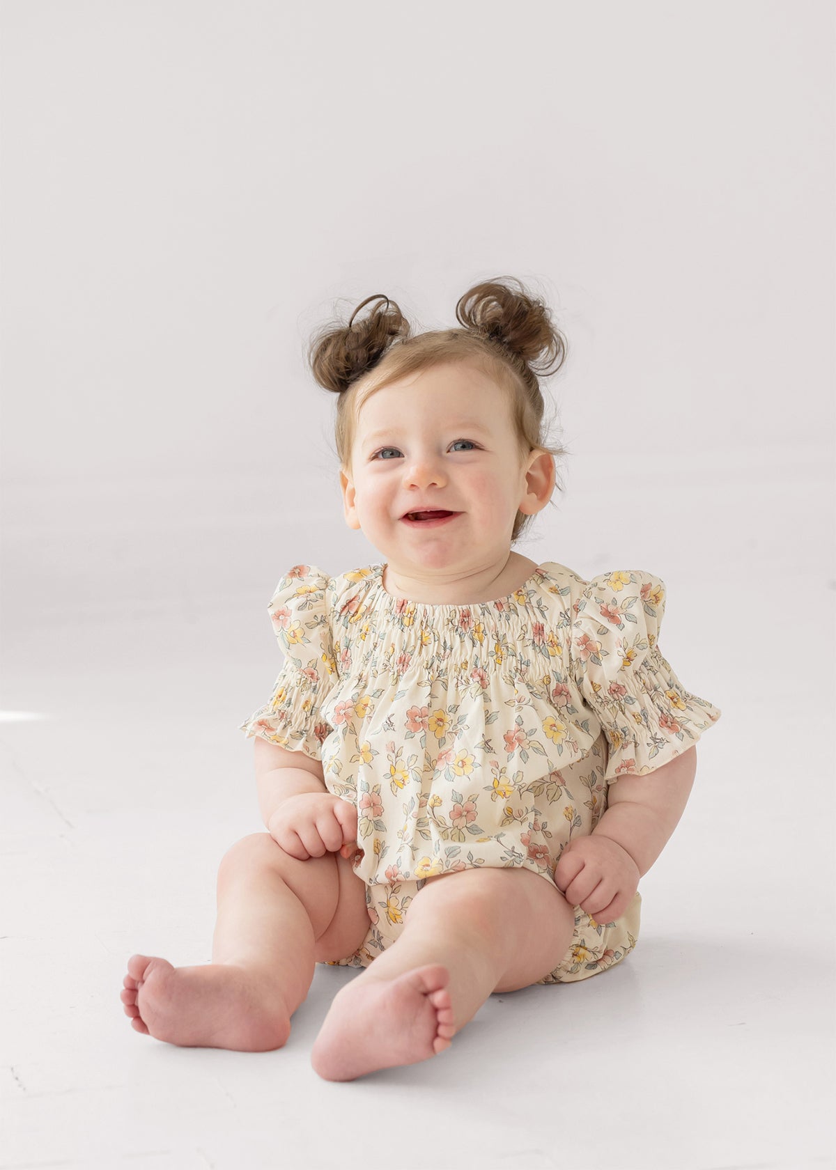 A smiling baby with light brown hair in two buns sits on the floor wearing the Mabel and Honey Audrey Flutter Sleeve Romper, featuring puffed sleeves and multicolored patterns, against a white background.