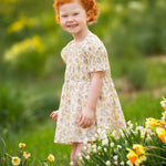 A young child with curly red hair smiles outdoors among yellow and white flowers, wearing the Audrey Flutter Sleeve Dress by Mabel and Honey. The blurred green background hints at a sunny spring or summer day.