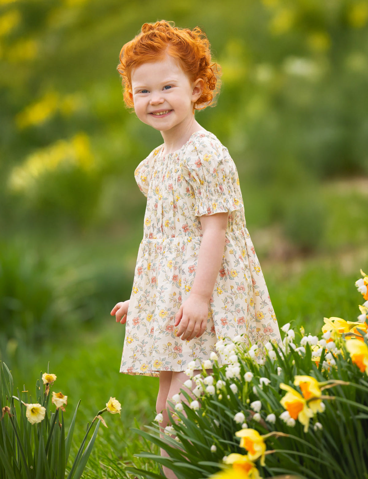 A young child with curly red hair smiles outdoors among yellow and white flowers, wearing the Audrey Flutter Sleeve Dress by Mabel and Honey. The blurred green background hints at a sunny spring or summer day.