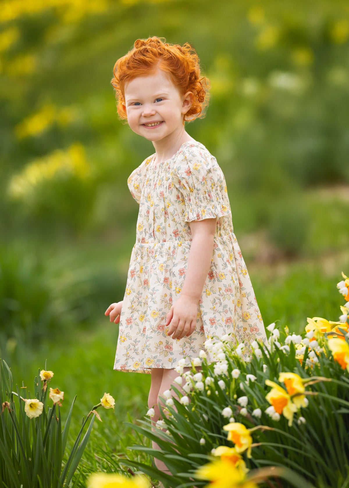 A young child with curly red hair smiles outdoors among yellow and white flowers, wearing the Audrey Flutter Sleeve Dress by Mabel and Honey. The blurred green background hints at a sunny spring or summer day.