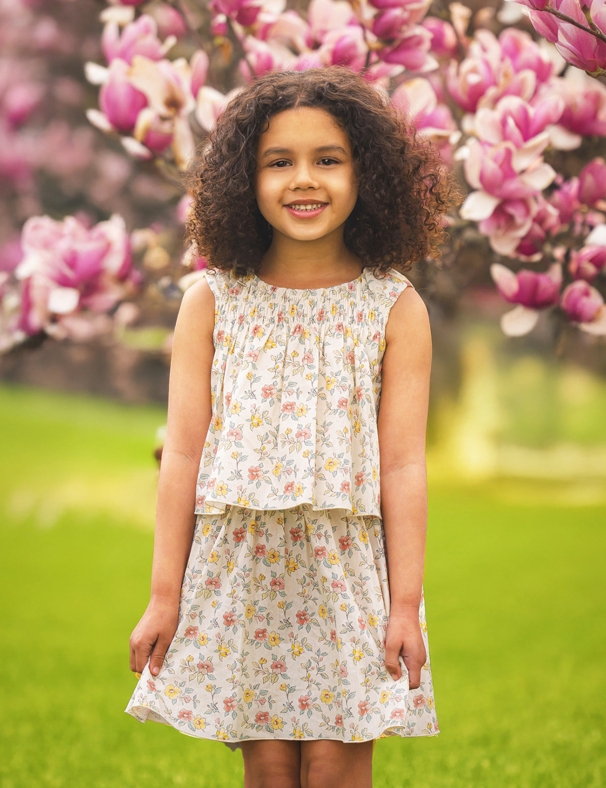 A young girl with curly hair and a bright smile wears the Audrey Sleeveless Dress by Mabel and Honey, standing outdoors among blooming pink magnolia flowers and green grass.