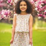 A young girl with curly hair and a bright smile wears the Audrey Sleeveless Dress by Mabel and Honey, standing outdoors among blooming pink magnolia flowers and green grass.