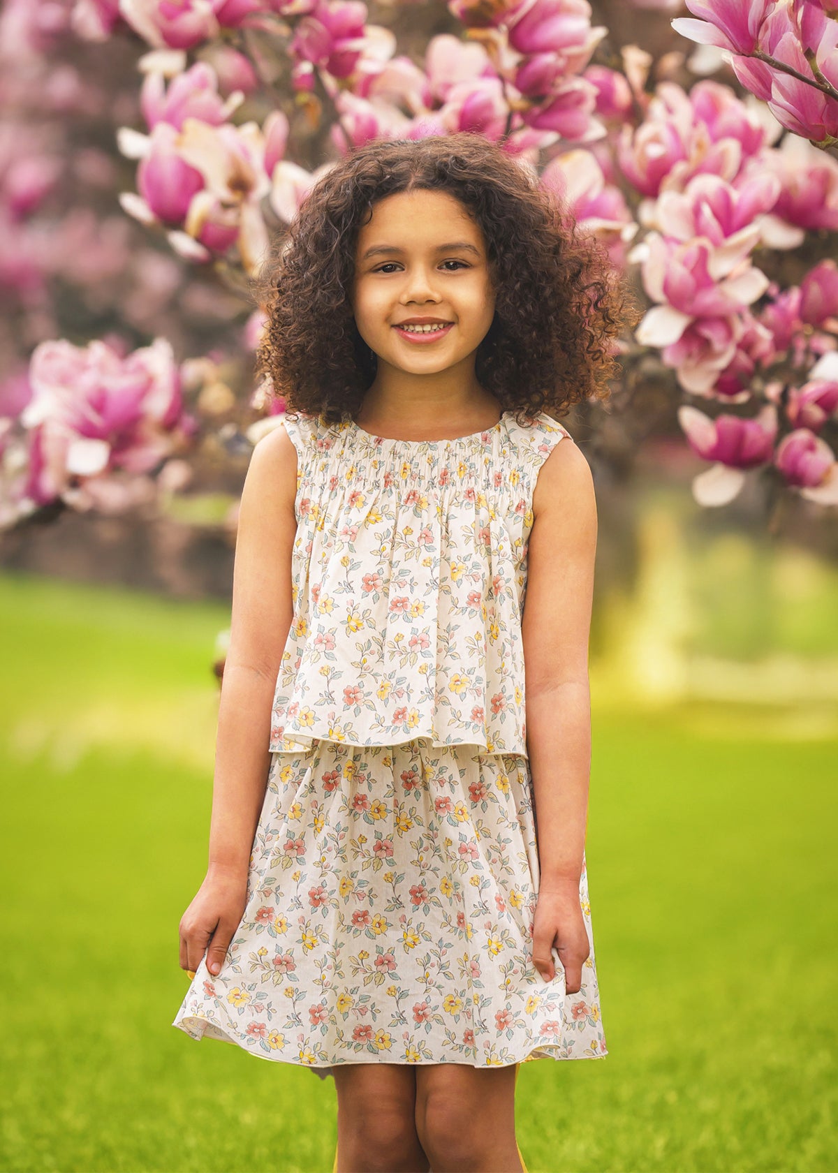 A young girl with curly hair and a bright smile wears the Audrey Sleeveless Dress by Mabel and Honey, standing outdoors among blooming pink magnolia flowers and green grass.