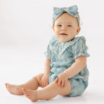 A baby wears the Mabel and Honey Delicate Blue Short Sleeve Romper with white embroidery and a matching headband, sitting on a white background and smiling slightly.
