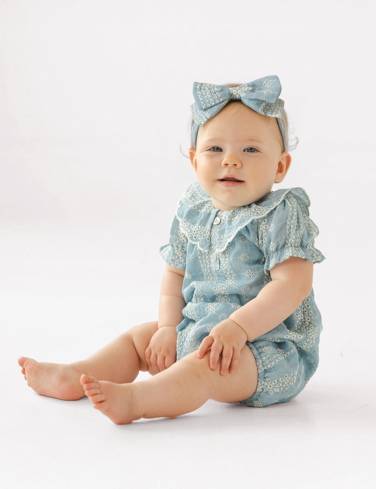 A baby wears the Mabel and Honey Delicate Blue Short Sleeve Romper with white embroidery and a matching headband, sitting on a white background and smiling slightly.