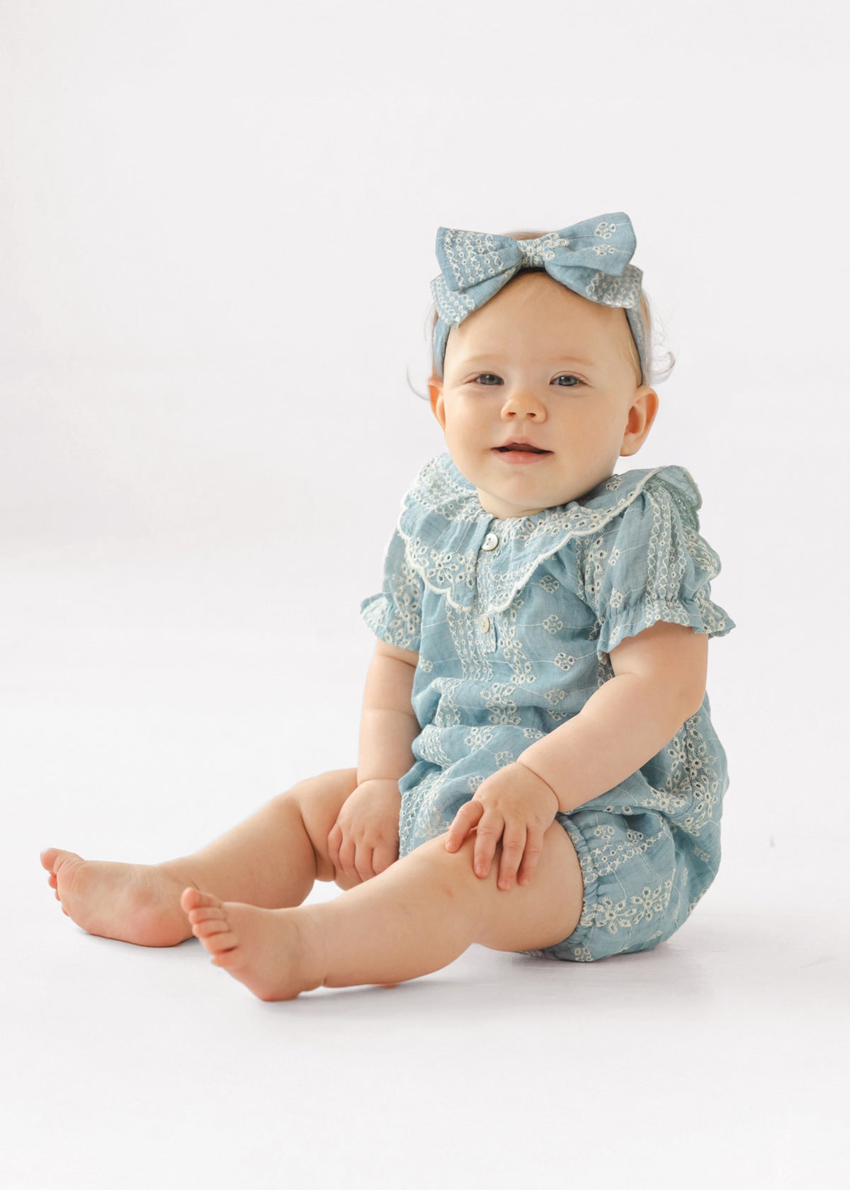 A baby wears the Mabel and Honey Delicate Blue Short Sleeve Romper with white embroidery and a matching headband, sitting on a white background and smiling slightly.