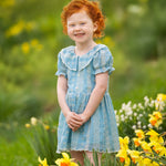 A young child with curly red hair smiles outdoors in a field of yellow daffodils, wearing the Mabel and Honey Delicate Blue Short Sleeve Dress. The green, blurred background suggests a bright spring day.