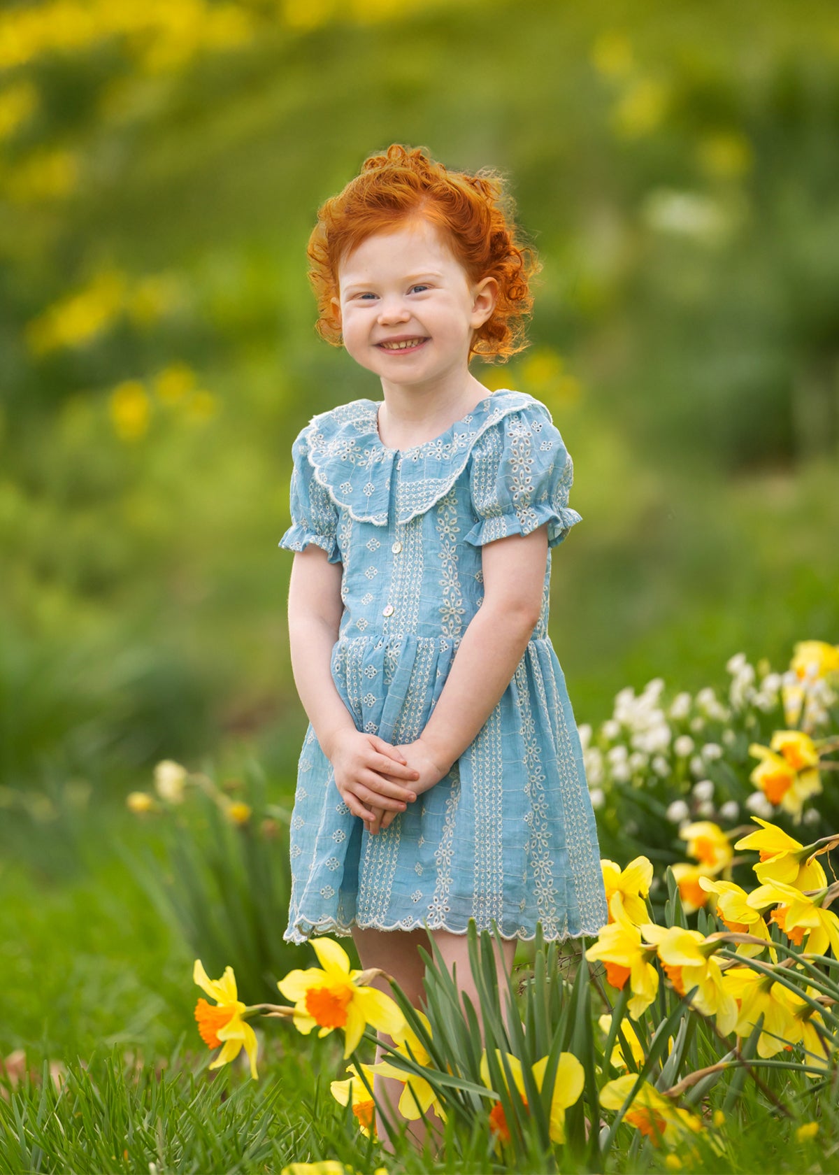 A young child with curly red hair smiles outdoors in a field of yellow daffodils, wearing the Mabel and Honey Delicate Blue Short Sleeve Dress. The green, blurred background suggests a bright spring day.