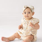 A baby wears the Mabel and Honey Confetti Cutie Cap Sleeve Romper with a matching bow headband, sitting on the floor against a plain white background.