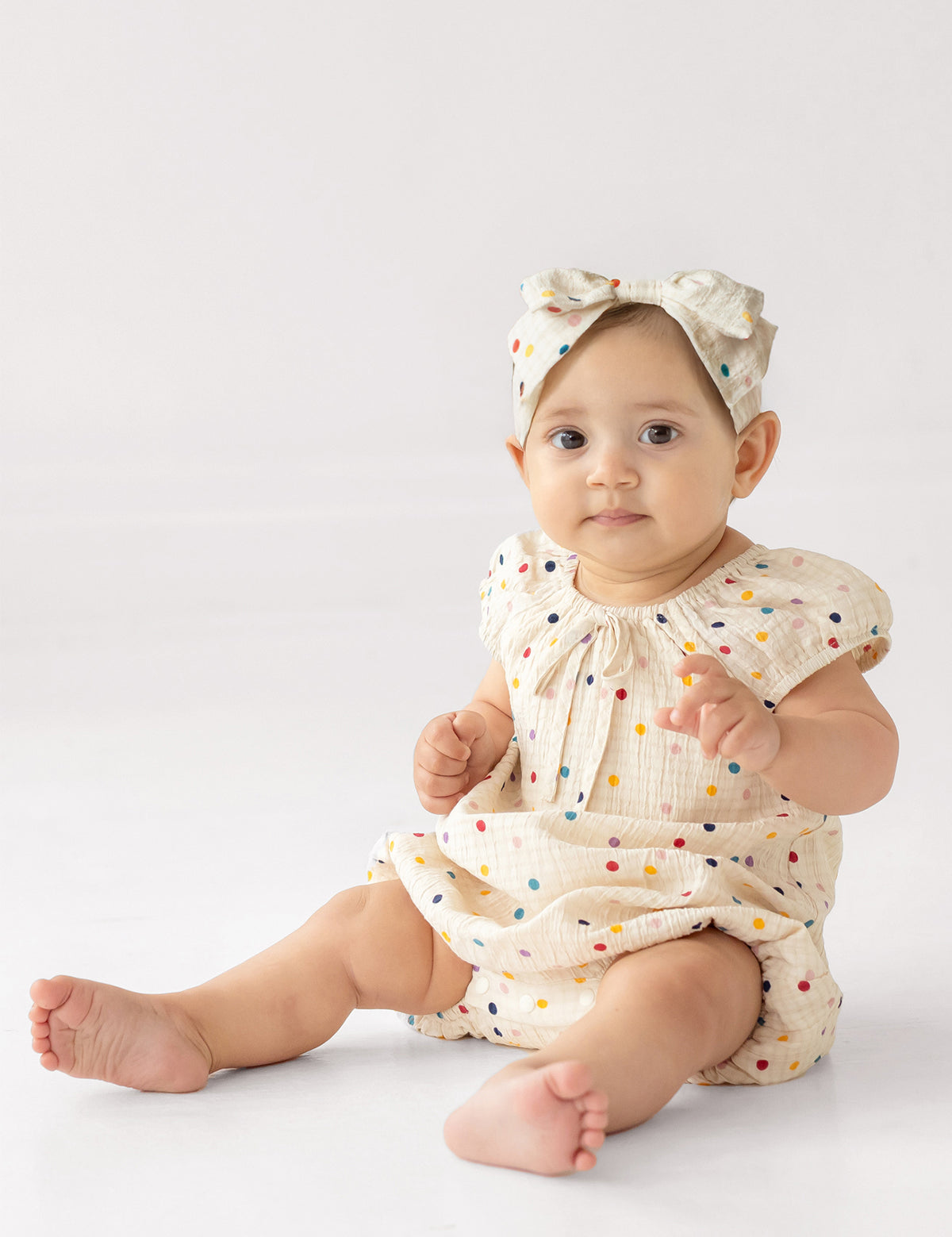 A baby wears the Mabel and Honey Confetti Cutie Cap Sleeve Romper with a matching bow headband, sitting on the floor against a plain white background.