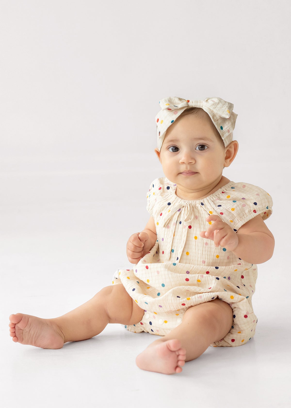 A baby wears the Mabel and Honey Confetti Cutie Cap Sleeve Romper with a matching bow headband, sitting on the floor against a plain white background.