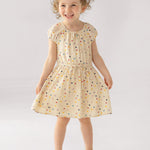 A young girl smiles in the Mabel and Honey Confetti Cutie Cap Sleeve Dress, featuring cream polka dots and a gathered waist. She pairs it with white sandals, posing against a simple light backdrop.