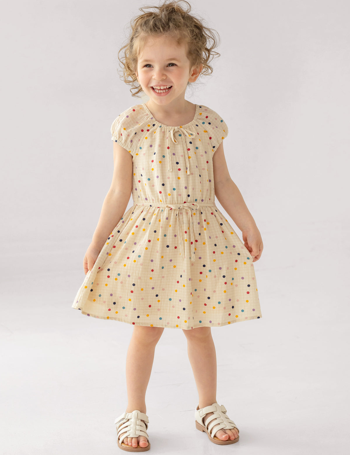 A young girl smiles in the Mabel and Honey Confetti Cutie Cap Sleeve Dress, featuring cream polka dots and a gathered waist. She pairs it with white sandals, posing against a simple light backdrop.