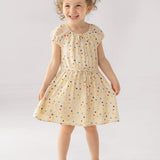 A young girl smiles in the Mabel and Honey Confetti Cutie Cap Sleeve Dress, featuring cream polka dots and a gathered waist. She pairs it with white sandals, posing against a simple light backdrop.