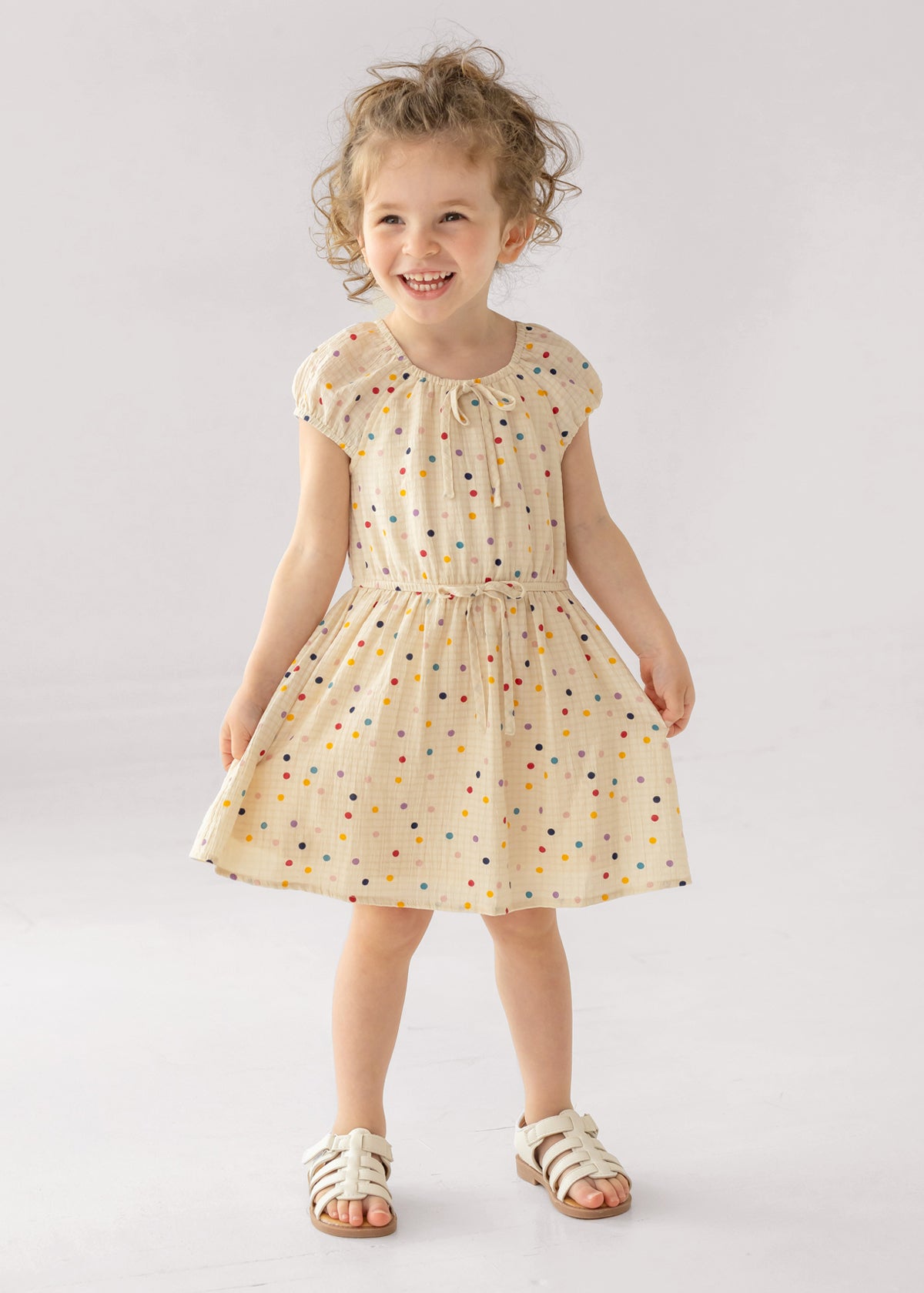 A young girl smiles in the Mabel and Honey Confetti Cutie Cap Sleeve Dress, featuring cream polka dots and a gathered waist. She pairs it with white sandals, posing against a simple light backdrop.