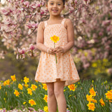 A young girl in the Tangerine Twirl Sleeveless Dress by Mabel and Honey stands barefoot among blooming daffodils, holding a yellow flower, with pink magnolia blossoms on trees in the background.
