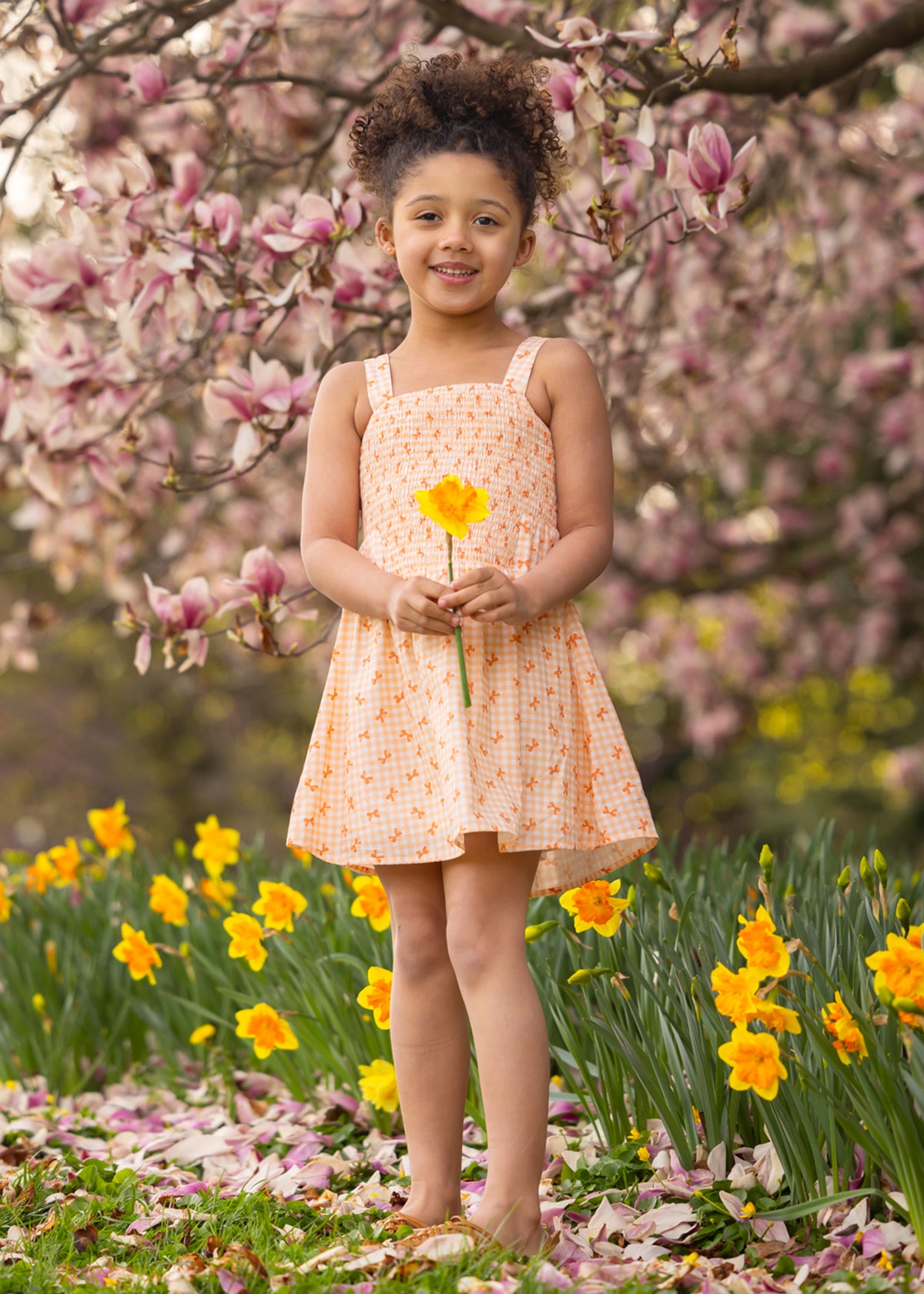 A young girl in the Tangerine Twirl Sleeveless Dress by Mabel and Honey stands barefoot among blooming daffodils, holding a yellow flower, with pink magnolia blossoms on trees in the background.