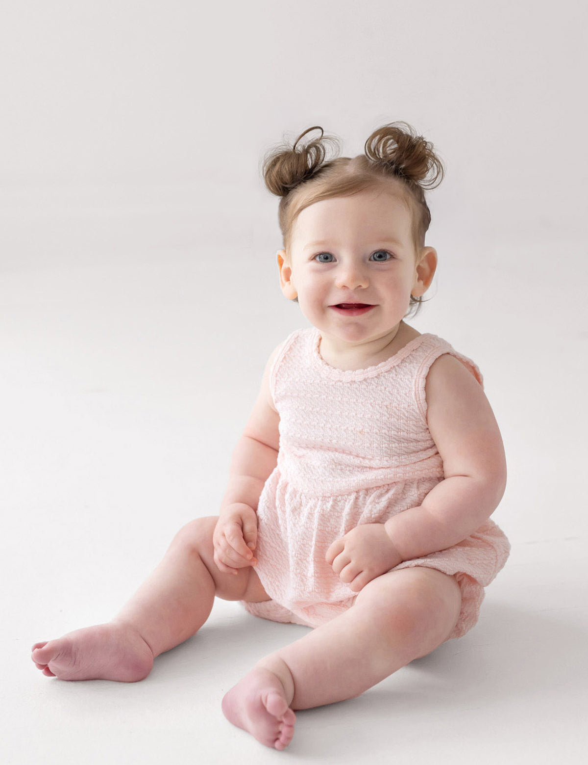 A smiling baby with blue eyes and light skin sits on the floor in the Mabel and Honey Pink Cloud Sleeveless Romper. Her light brown hair is styled in two small buns, and the background is plain white.