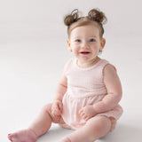 A smiling baby with blue eyes and light skin sits on the floor in the Mabel and Honey Pink Cloud Sleeveless Romper. Her light brown hair is styled in two small buns, and the background is plain white.