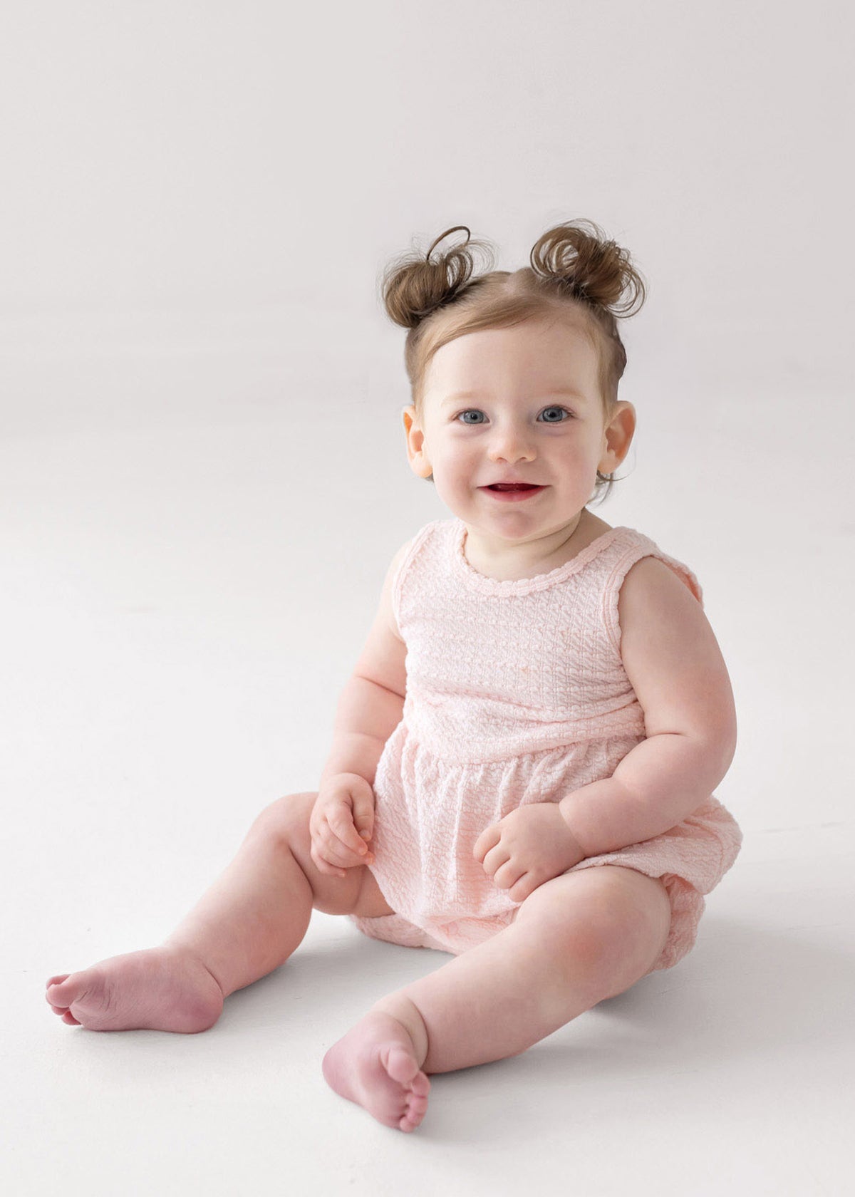 A smiling baby with blue eyes and light skin sits on the floor in the Mabel and Honey Pink Cloud Sleeveless Romper. Her light brown hair is styled in two small buns, and the background is plain white.