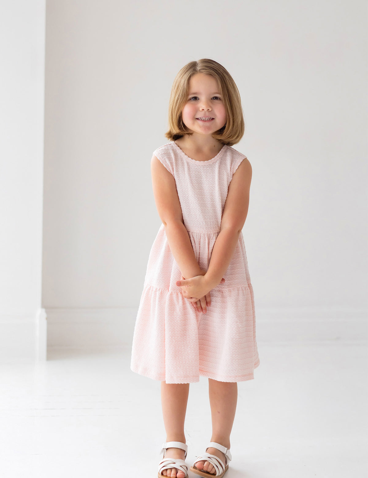A girl in the Mabel and Honey Pink Cloud Sleeveless Dress, featuring a tiered gathered skirt and a charming back bow detail.