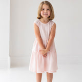 A girl in the Mabel and Honey Pink Cloud Sleeveless Dress, featuring a tiered gathered skirt and a charming back bow detail.