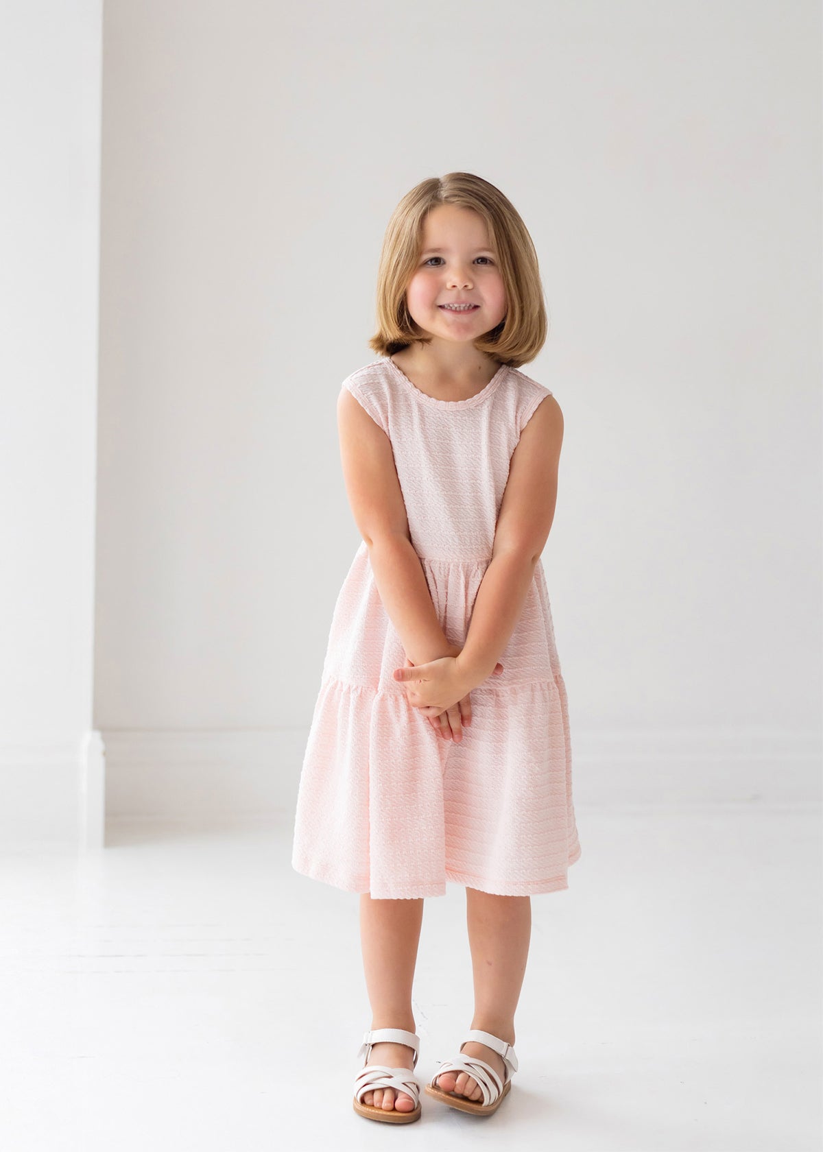 A girl in the Mabel and Honey Pink Cloud Sleeveless Dress, featuring a tiered gathered skirt and a charming back bow detail.