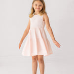 A young girl in the Mabel and Honey Pink Cloud Sleeveless Dress stands with arms slightly outstretched, smiling in translucent sandals against a plain white background.