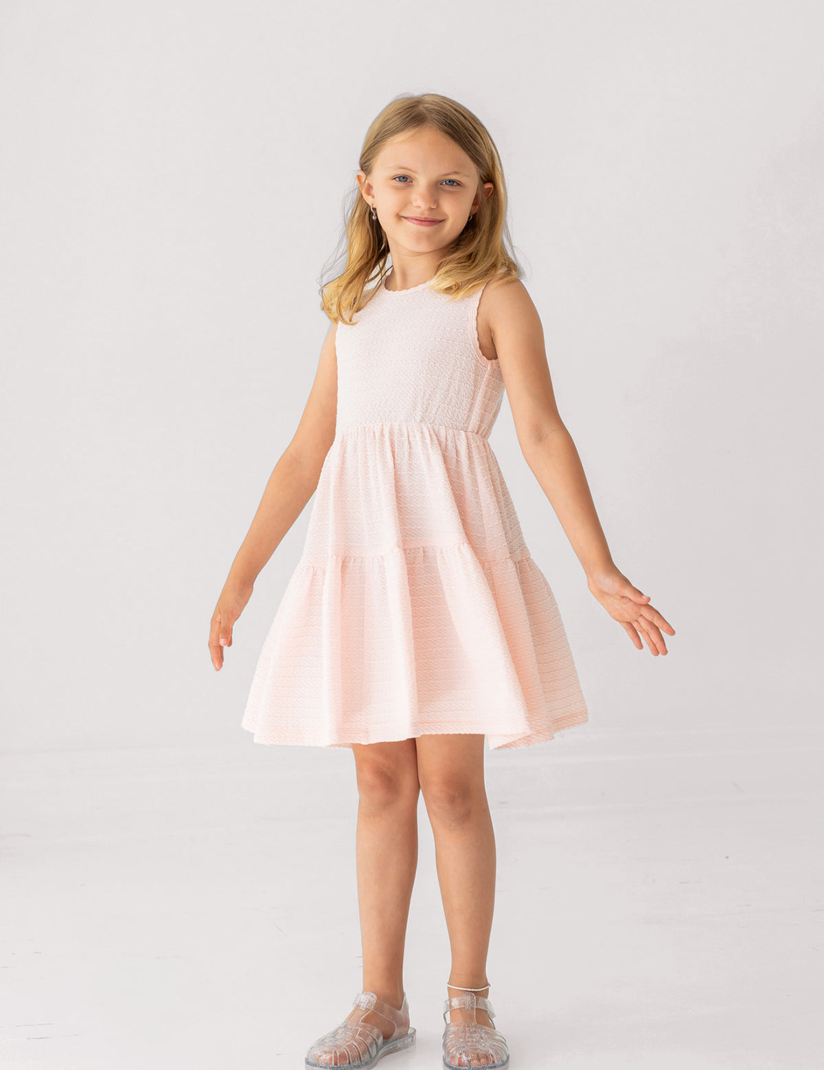 A young girl in the Mabel and Honey Pink Cloud Sleeveless Dress stands with arms slightly outstretched, smiling in translucent sandals against a plain white background.