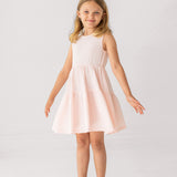 A young girl in the Mabel and Honey Pink Cloud Sleeveless Dress stands with arms slightly outstretched, smiling in translucent sandals against a plain white background.