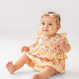 A baby sits on the floor wearing the Mabel and Honey Blooming Florals Short Gathered Sleeve Romper with a matching headband, smiling slightly while looking forward against a simple light background.
