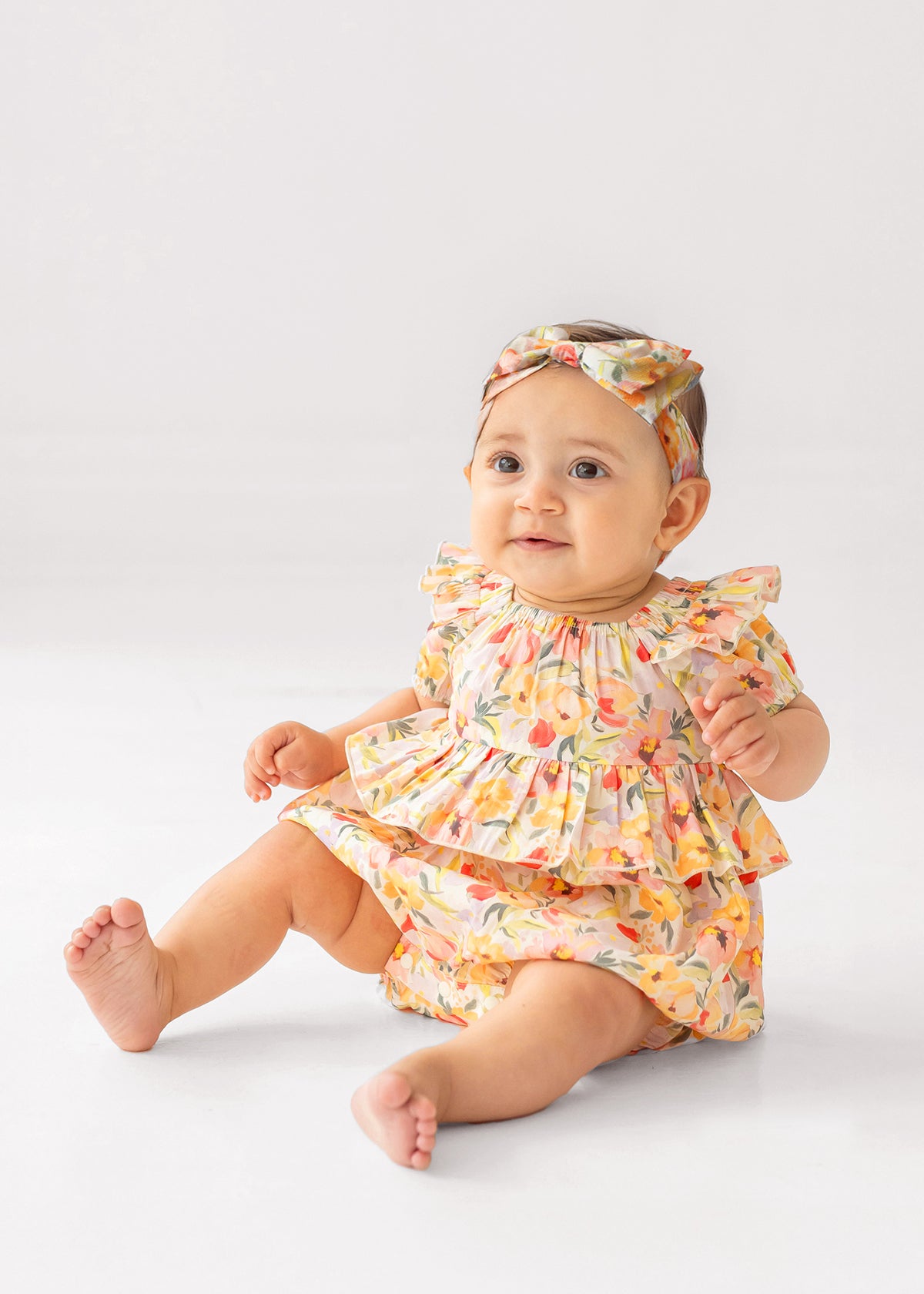 A baby sits on the floor wearing the Mabel and Honey Blooming Florals Short Gathered Sleeve Romper with a matching headband, smiling slightly while looking forward against a simple light background.