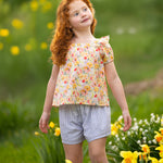 A young girl with curly red hair smiles gently among yellow and white flowers, wearing the Blooming Florals Short Gathered Sleeve Two Piece Set by Mabel and Honey, set against a softly blurred green background.