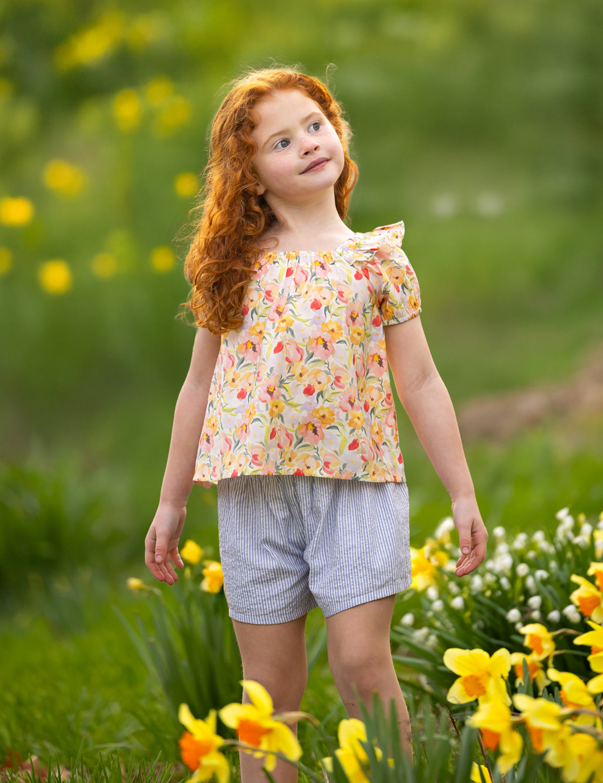 A young girl with curly red hair smiles gently among yellow and white flowers, wearing the Blooming Florals Short Gathered Sleeve Two Piece Set by Mabel and Honey, set against a softly blurred green background.
