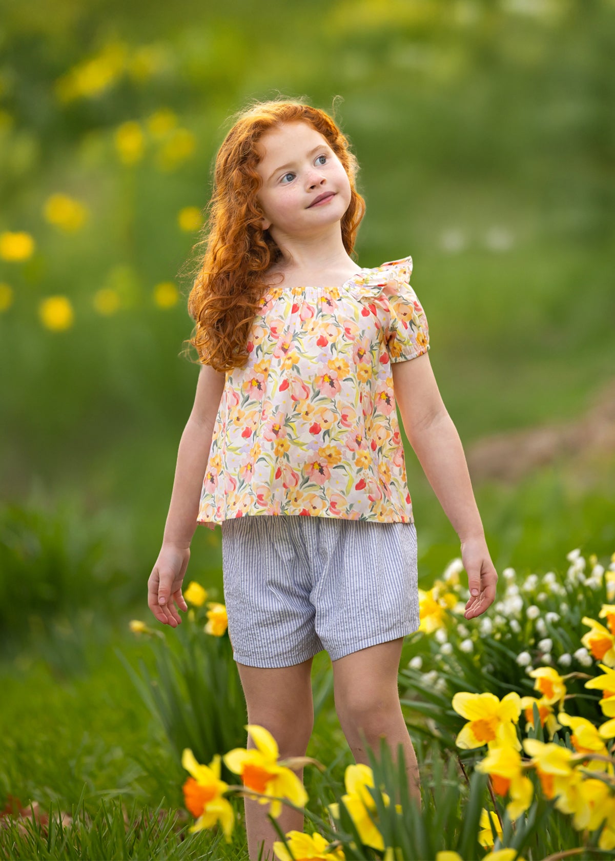 A young girl with curly red hair smiles gently among yellow and white flowers, wearing the Blooming Florals Short Gathered Sleeve Two Piece Set by Mabel and Honey, set against a softly blurred green background.