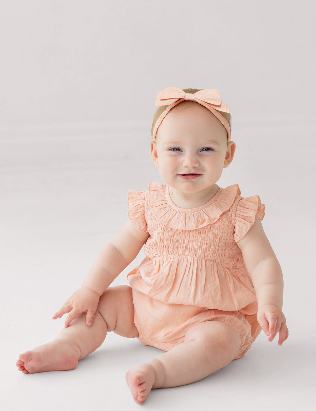 A smiling baby wearing the Mabel and Honey Jane Ruffled Short Sleeve Romper in peach, paired with a matching bow headband, sits barefoot on a light, neutral background.