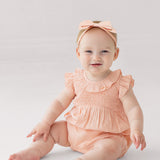A smiling baby wearing the Mabel and Honey Jane Ruffled Short Sleeve Romper in peach, paired with a matching bow headband, sits barefoot on a light, neutral background.