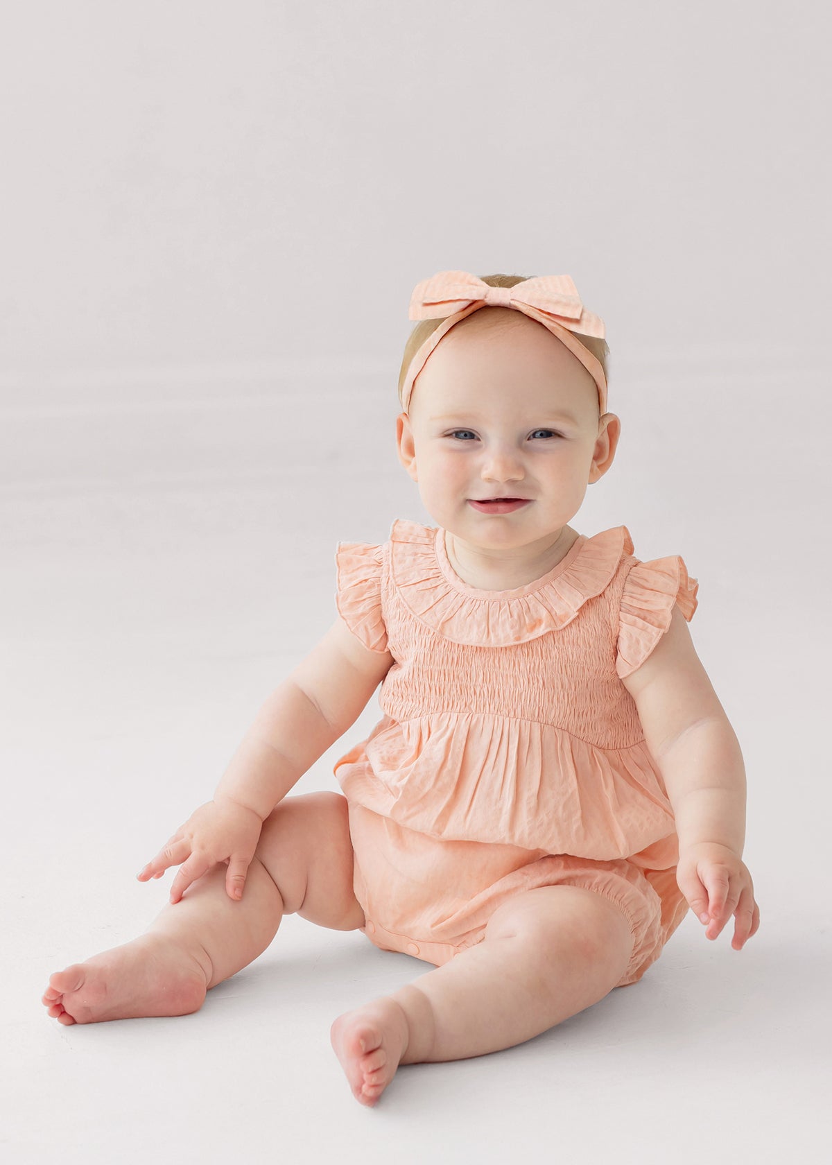 A smiling baby wearing the Mabel and Honey Jane Ruffled Short Sleeve Romper in peach, paired with a matching bow headband, sits barefoot on a light, neutral background.