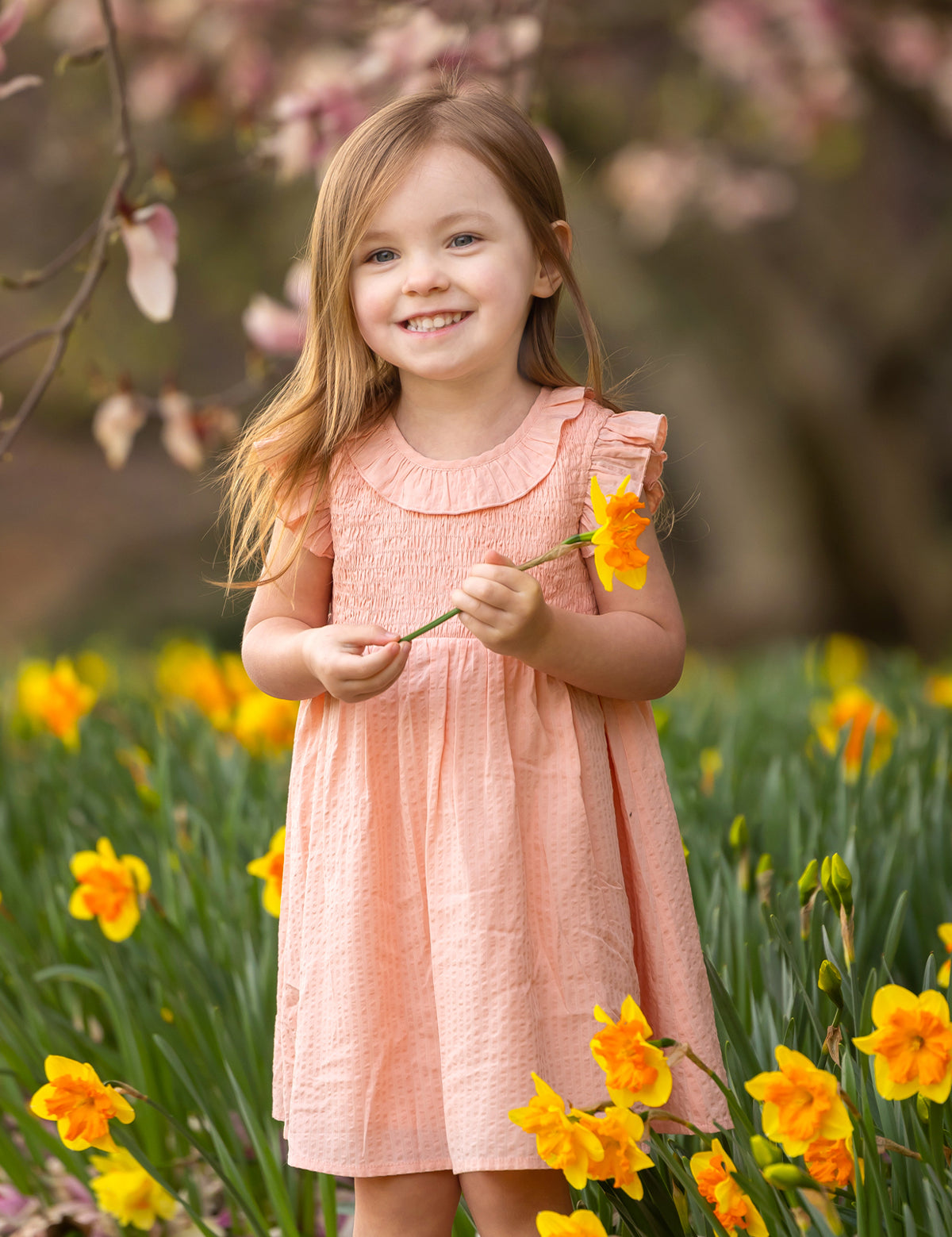 A young girl smiles in a field of yellow daffodils, wearing the Mabel and Honey Jane Ruffled Short Sleeve Dress. She holds a daffodil, with blurred pink blossoms and green foliage in the background.