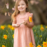 A young girl smiles in a field of yellow daffodils, wearing the Mabel and Honey Jane Ruffled Short Sleeve Dress. She holds a daffodil, with blurred pink blossoms and green foliage in the background.