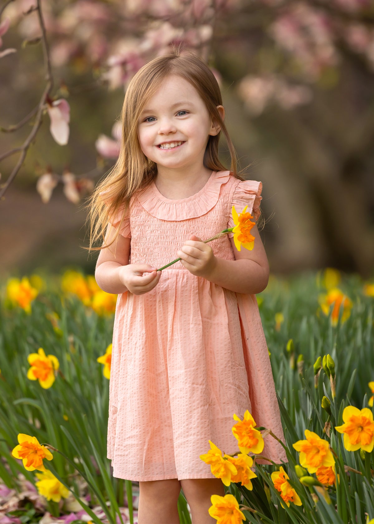 A young girl smiles in a field of yellow daffodils, wearing the Mabel and Honey Jane Ruffled Short Sleeve Dress. She holds a daffodil, with blurred pink blossoms and green foliage in the background.