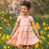 A young girl smiles among yellow daffodils, wearing the Jane Tank Dress by Mabel and Honey. Light peach with an A-line silhouette, her dress stands out against a backdrop of blooming pink magnolia blossoms, capturing a vibrant spring scene.