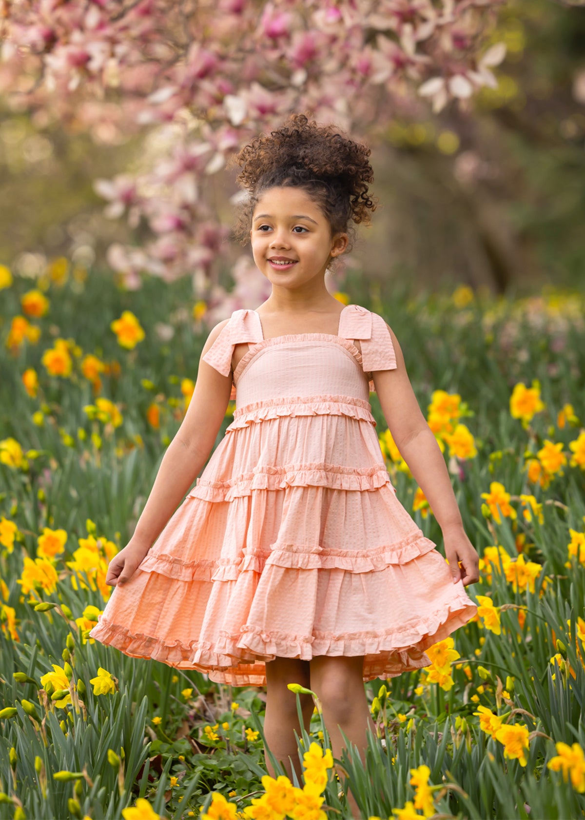 A young girl smiles among yellow daffodils, wearing the Jane Tank Dress by Mabel and Honey. Light peach with an A-line silhouette, her dress stands out against a backdrop of blooming pink magnolia blossoms, capturing a vibrant spring scene.