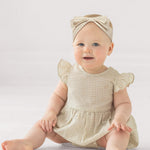 A baby in the Mabel and Honey Dakota Ruffled Short Sleeve Romper, featuring a beige and white checkered pattern with ruffled sleeves and a matching headband, sits smiling on the floor against a plain light background.