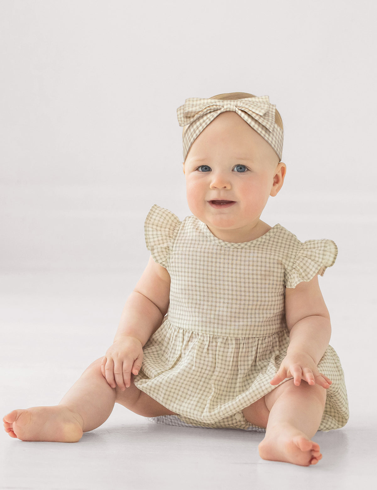 A baby in the Mabel and Honey Dakota Ruffled Short Sleeve Romper, featuring a beige and white checkered pattern with ruffled sleeves and a matching headband, sits smiling on the floor against a plain light background.