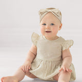 A baby in the Mabel and Honey Dakota Ruffled Short Sleeve Romper, featuring a beige and white checkered pattern with ruffled sleeves and a matching headband, sits smiling on the floor against a plain light background.